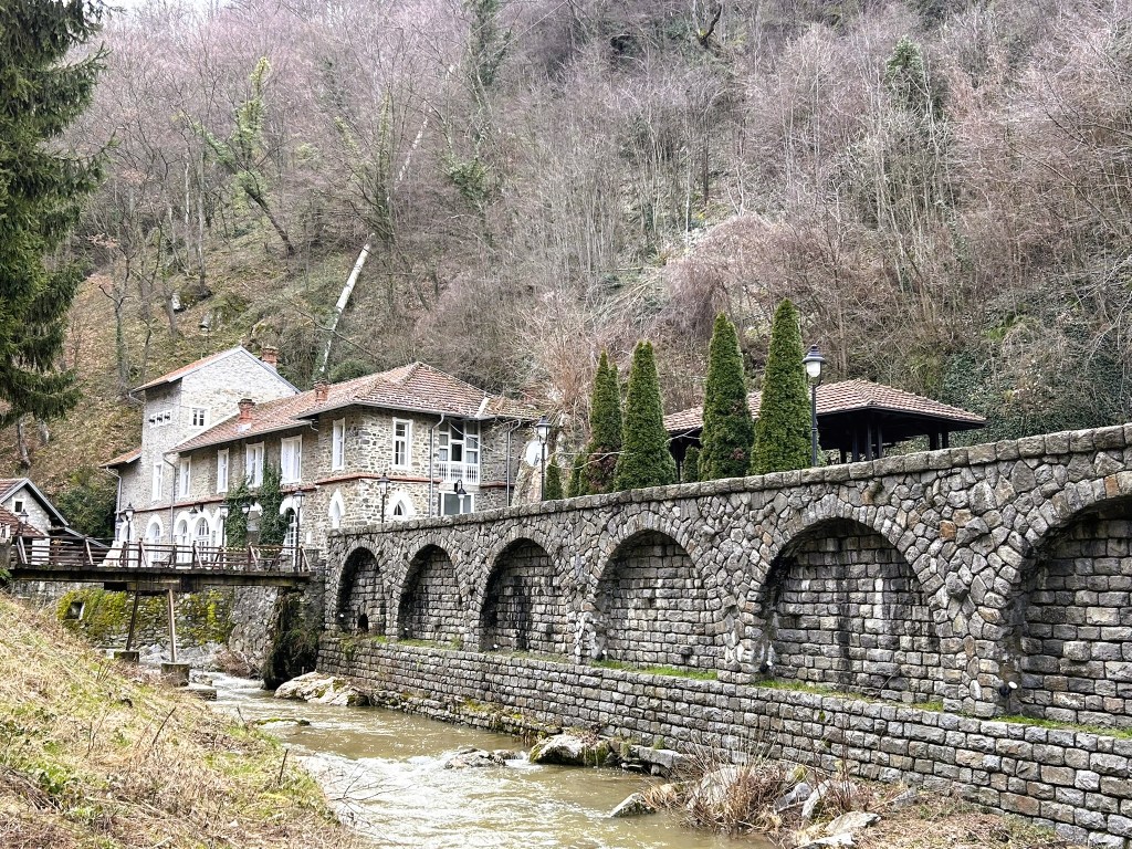 A stone wall with arches next to a flowing river, with a historic building and tall trees in the background surrounded by a hillside.
