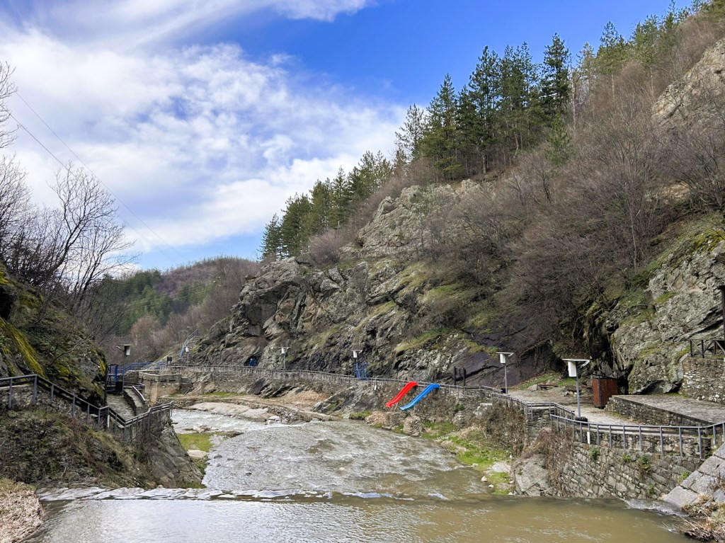 A scenic view of a river running through rocky terrain, flanked by trees and a walking path. The landscape features a small slide in red and blue, adding a playful element to the natural setting.