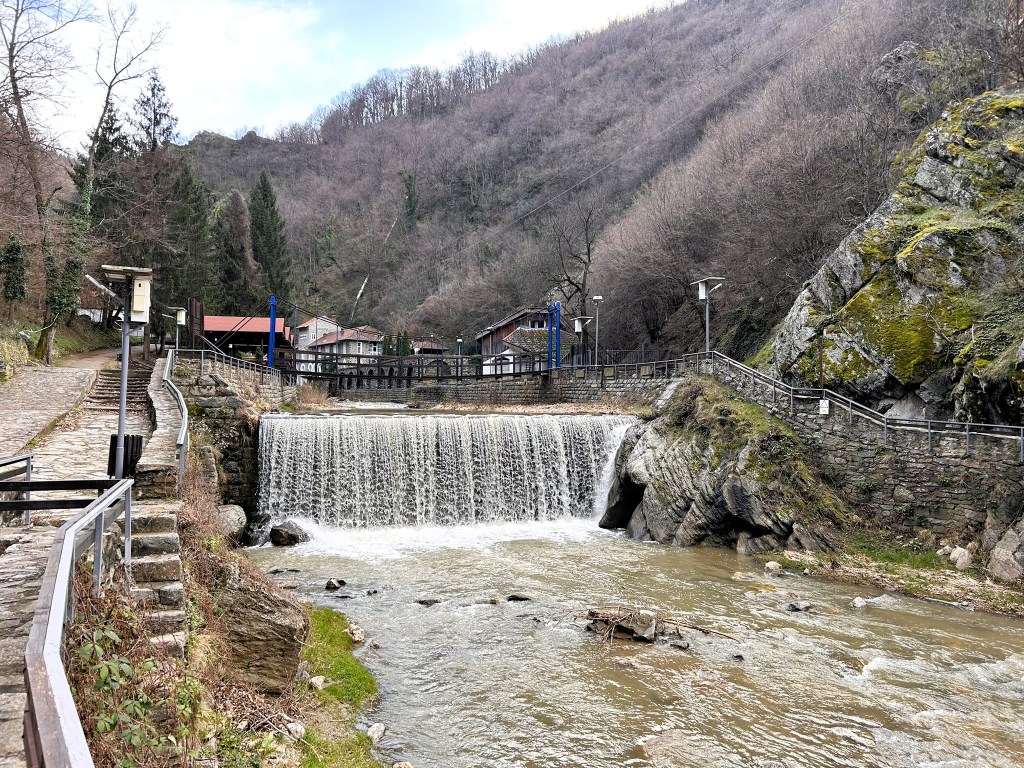 A scenic view of a waterfall cascading over rocks, surrounded by trees and hills. A path leads alongside the water, with rustic buildings visible in the background.