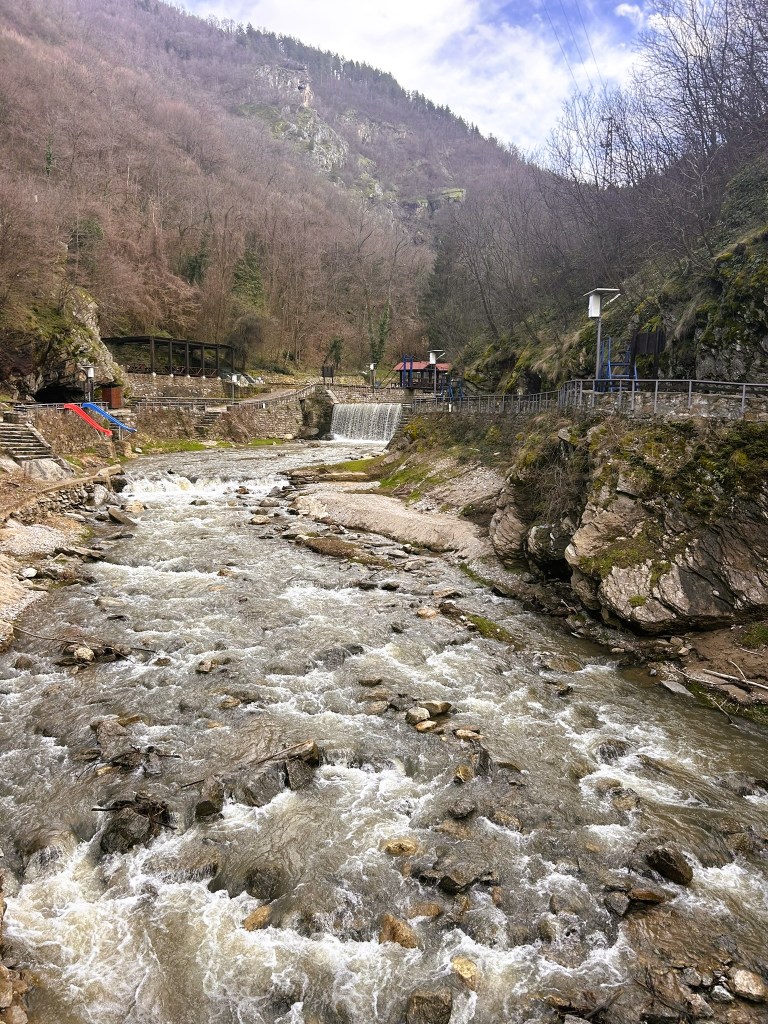 A peaceful river flowing through a rocky landscape, with a small waterfall and greenery on the hillsides, under a cloudy sky.