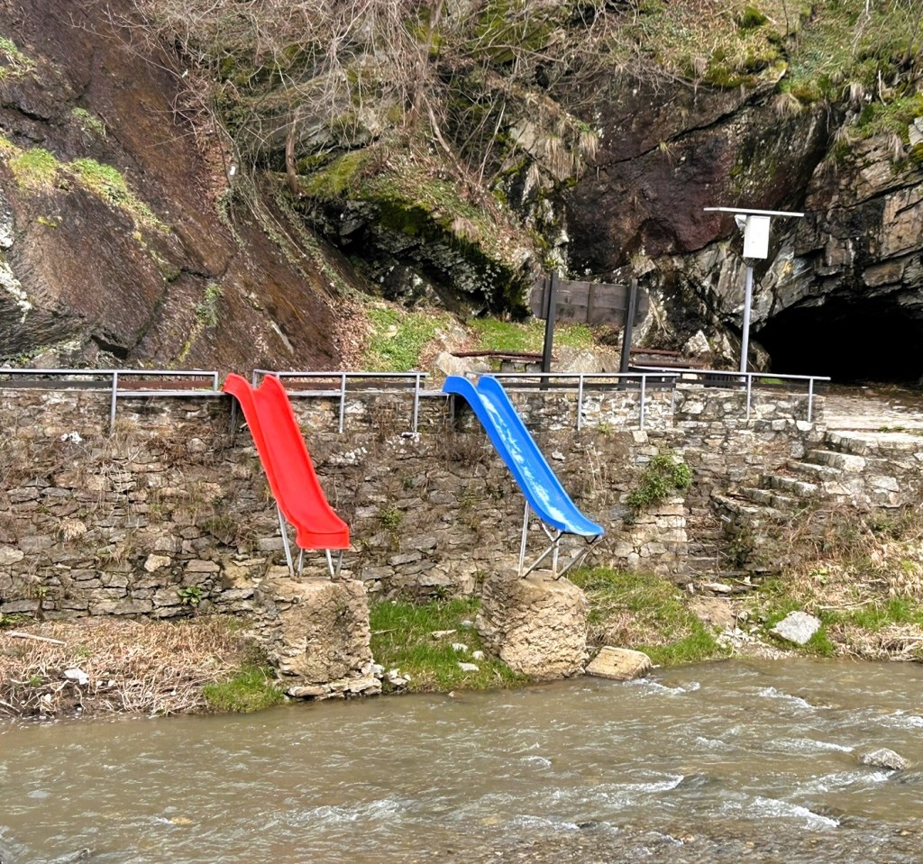 Two water slides, one red and one blue, positioned on stone supports beside a river, with rocky terrain and vegetation in the background.