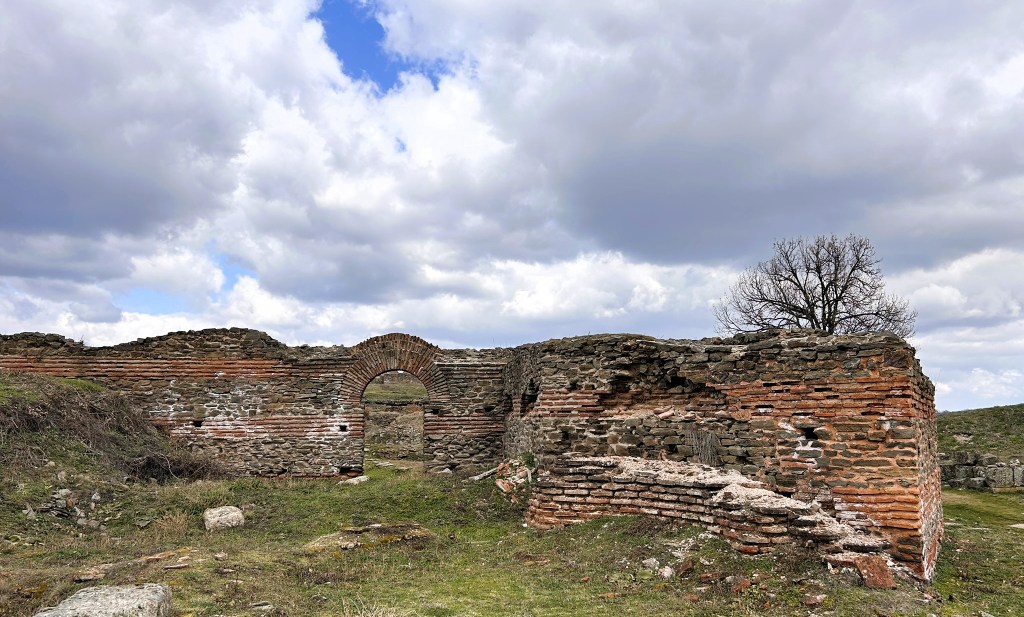 Ruins of an ancient brick structure with an arched opening, surrounded by grass and rocky terrain under a cloudy sky.