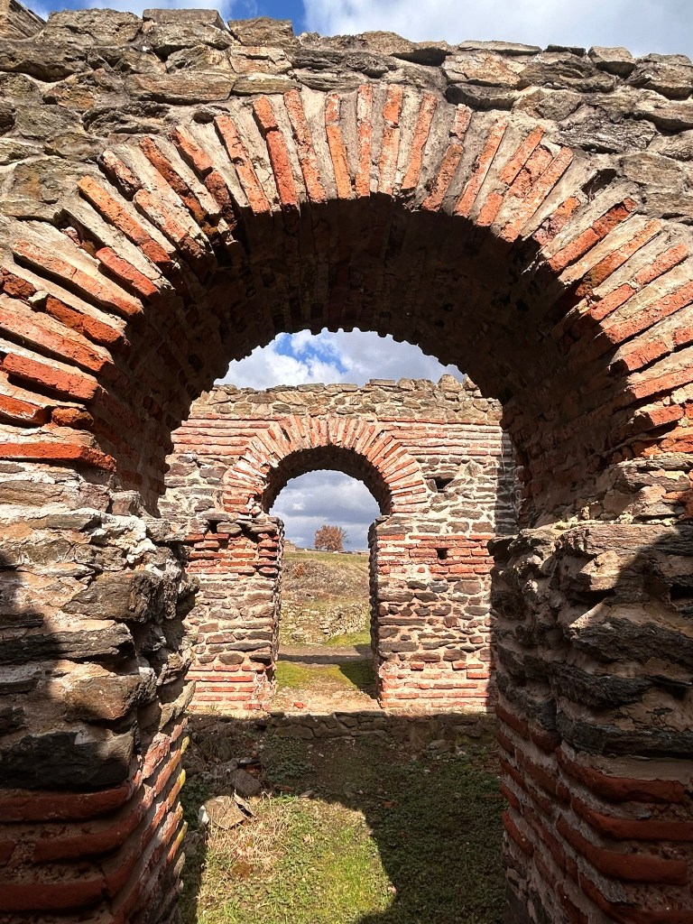 View of ancient stone ruins featuring arched brickwork, with a grassy area beyond and cloudy skies in the background.