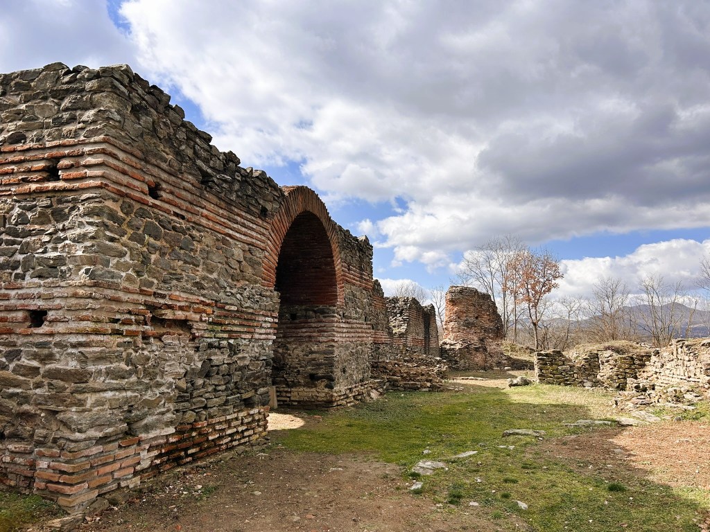 Ancient stone ruins with brick detailing set against a cloudy sky, featuring overgrown grass and trees in the background.