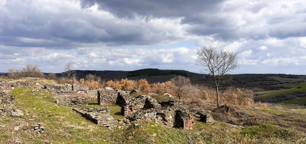 Ruins of an ancient stone structure on a grassy hill, with a backdrop of rolling hills and a cloudy sky.