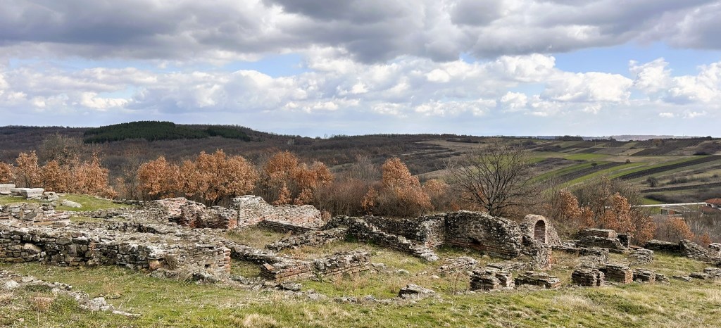 View of ancient stone ruins on a hillside, surrounded by autumn trees and rolling green fields under a cloudy sky.