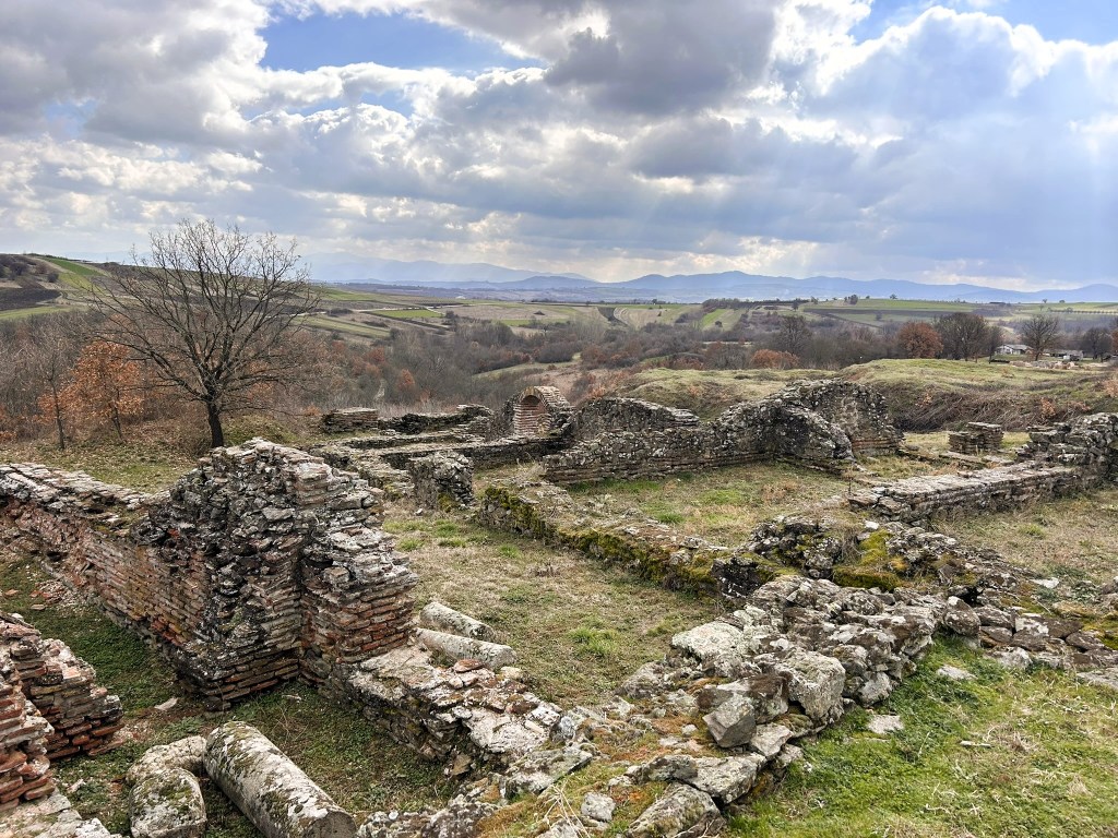 Ruins of ancient stone structures in a grassy landscape, surrounded by trees and hills under a cloudy sky.
