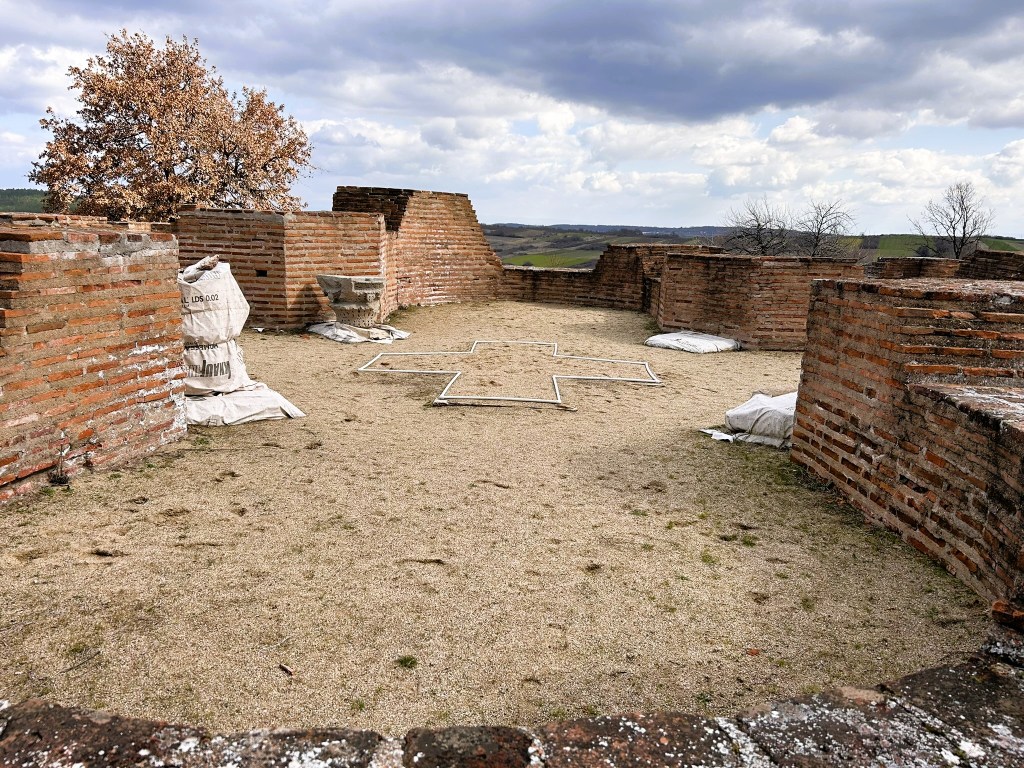 Ruins of an ancient building surrounded by brick walls, with a sandy floor and scattered bags on the ground. A tree is visible in the background under a cloudy sky.