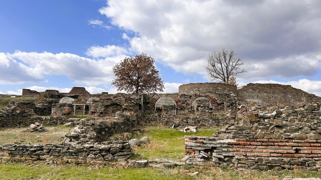 Ruins of an ancient structure surrounded by grassy areas and trees under a partly cloudy sky.