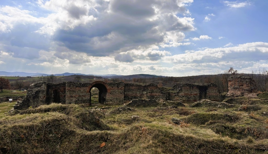 Ruins of an ancient stone structure surrounded by grassy fields and hills under a cloudy sky.