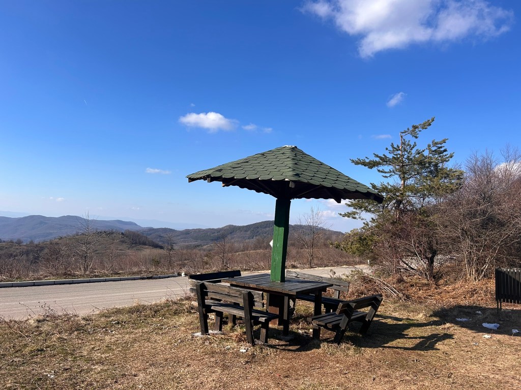 A wooden picnic table and benches under a green-roofed gazebo, set against a backdrop of rolling hills and a clear blue sky.