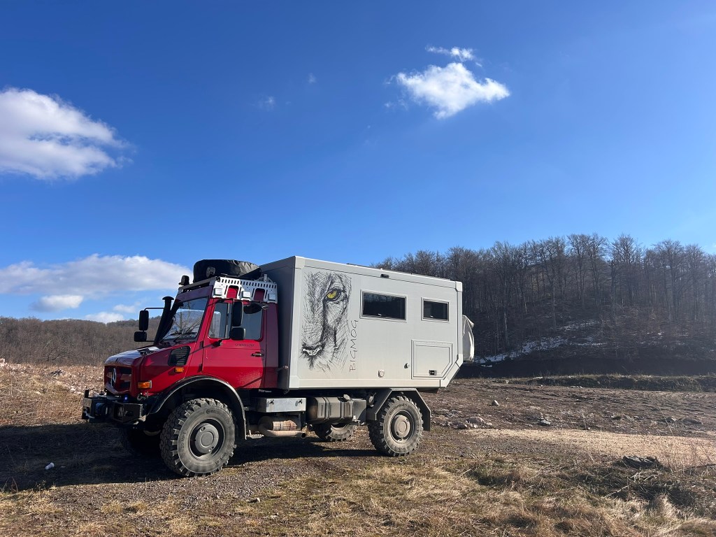 A red off-road vehicle with a grey camper body, featuring a lion's eye mural, parked in a rural landscape with sparse vegetation and a distant forest under a clear blue sky.
