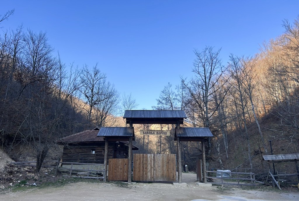 Entrance gate to a forested area with a rustic wooden structure beside it, under a clear blue sky.
