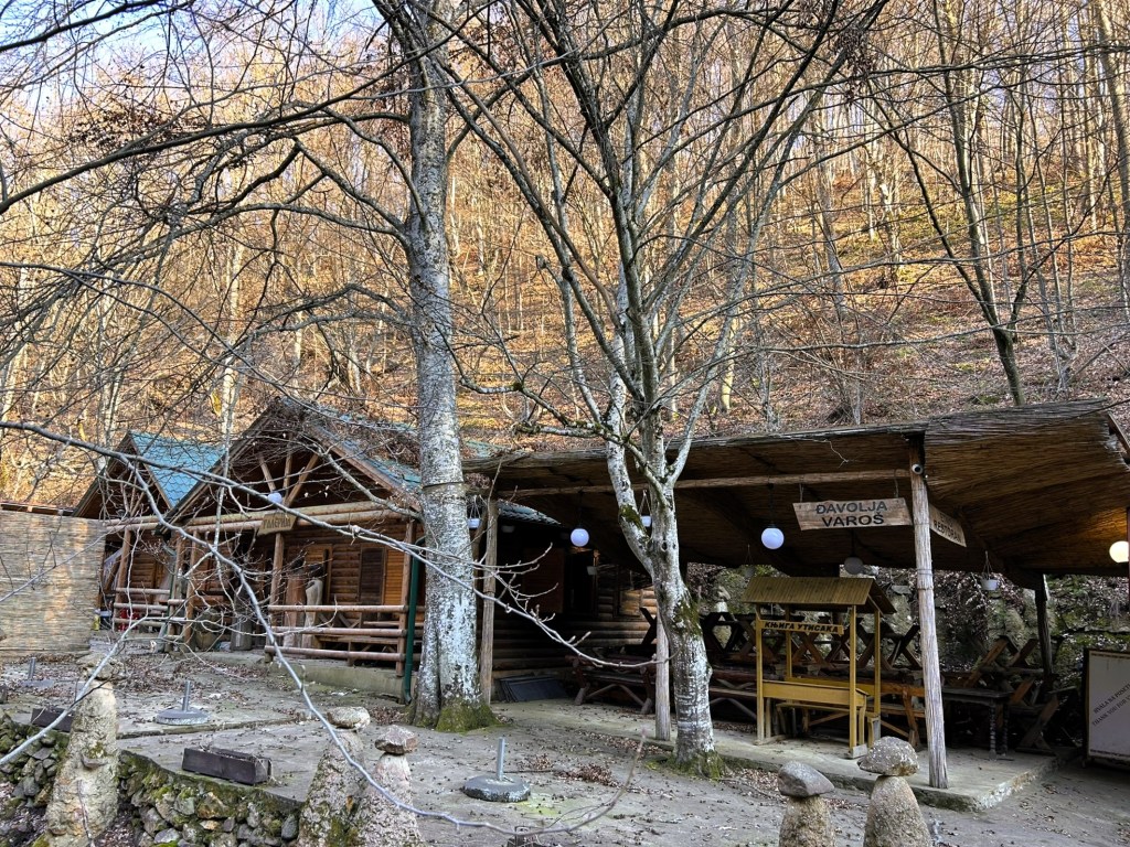 A wooden cabin surrounded by bare trees on a hillside during autumn. The cabin features a sloped roof and is partially shaded, with wooden signage visible. Stone features are placed in the foreground, adding to the rustic charm of the scene.