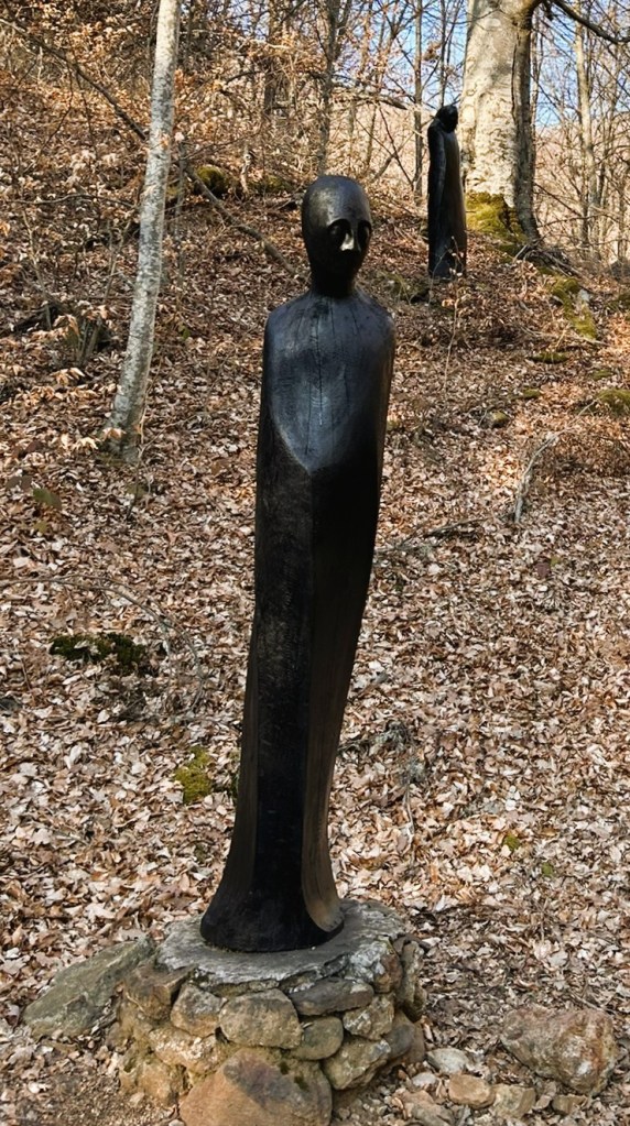 A close-up view of a tall, black wooden sculpture resembling a humanoid figure, standing on a stone base in a forest with dry leaves and trees in the background.