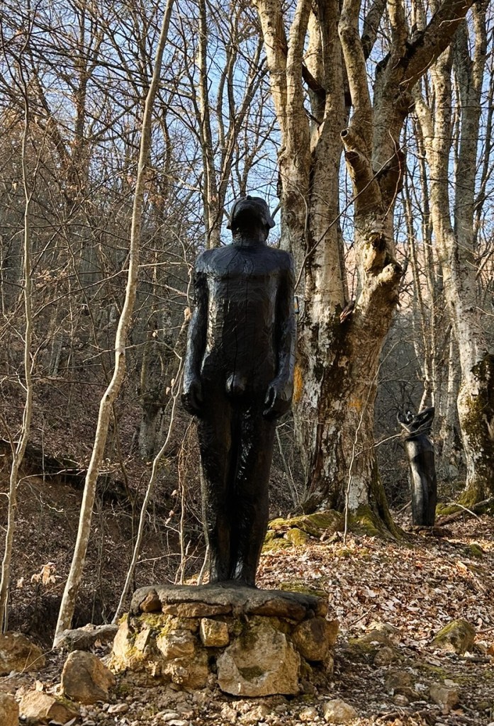 A black sculpture of a standing figure on a stone base, surrounded by bare trees and fallen leaves in a forested area.