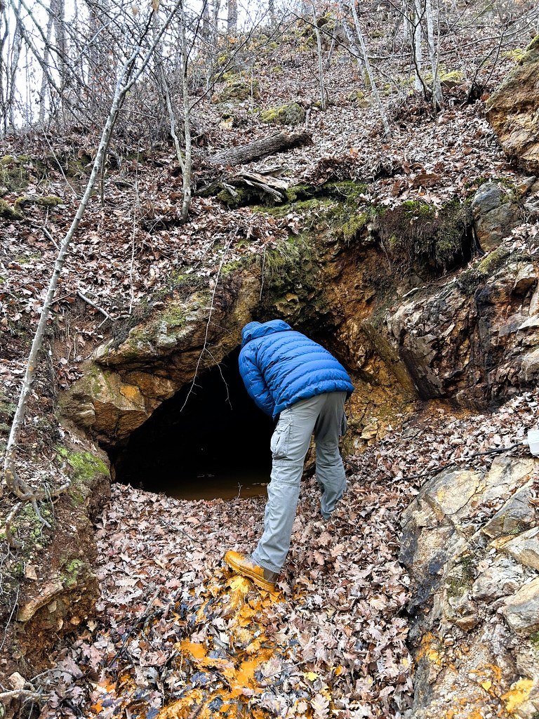 A person in a blue jacket leans into a dark cave entrance, surrounded by rocky terrain and fallen leaves.