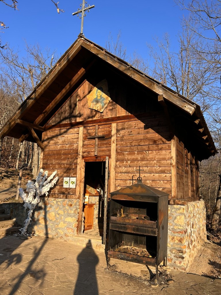 A small wooden chapel featuring a cross on the roof and a religious painting above the entrance, surrounded by bare trees and a stone path.