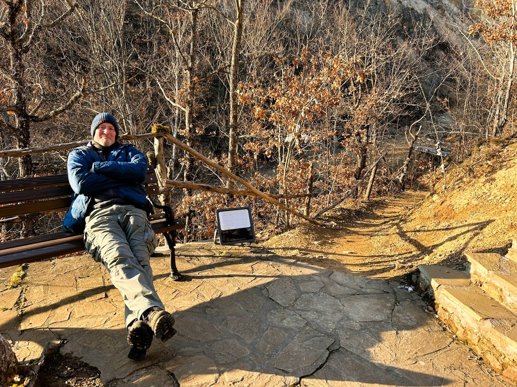 A person sitting on a bench, wearing a blue jacket and a beanie, in a forested area with bare trees and a stone pathway leading into the distance.