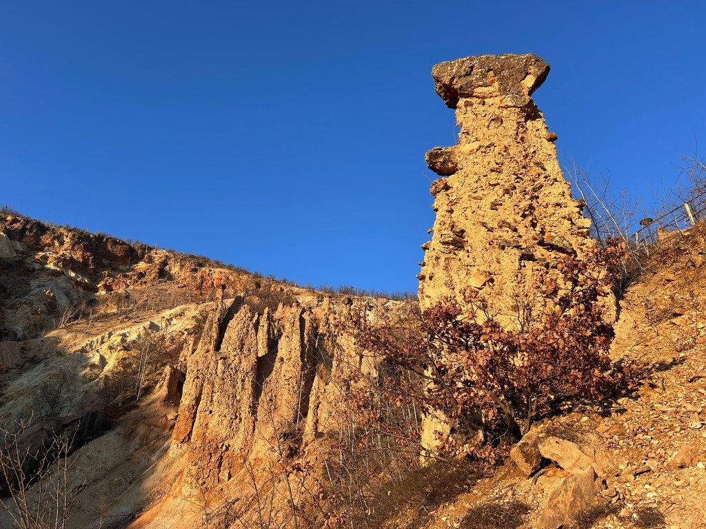 A tall, weathered rock formation stands prominently against a clear blue sky, surrounded by steep, rocky terrain and sparse vegetation.