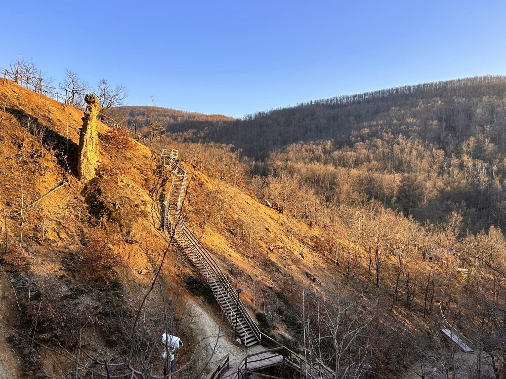 A wooden staircase winding up a steep hill, surrounded by sparse trees and a hilly landscape under a clear sky.
