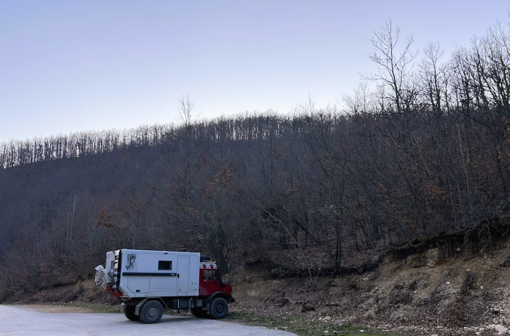 A rugged truck parked on a roadside near a forested hillside with bare trees, under a clear sky.