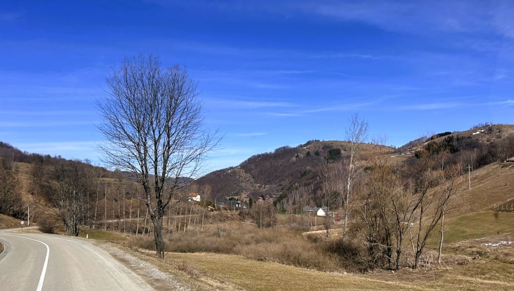 A winding road through a rural landscape featuring leafless trees, rolling hills, and a clear blue sky.