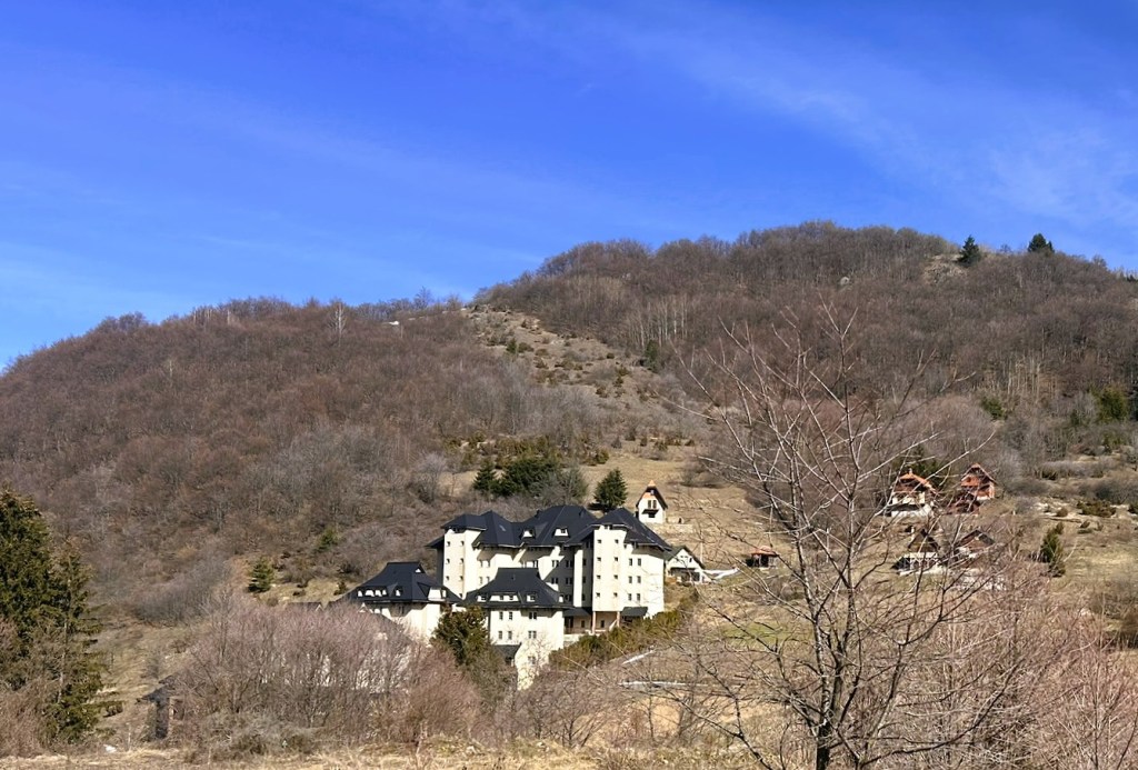 A large building with a dark roof situated on a hillside surrounded by bare trees and a mountainous landscape under a clear blue sky.