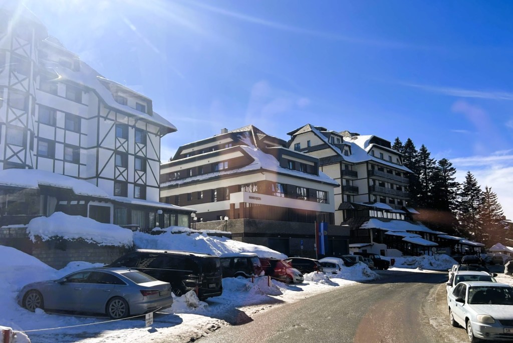 Snow-covered buildings with a mix of architectural styles, including modern and traditional, set against a clear blue sky. The scene features a road lined with parked cars and snowdrifts, indicating a winter setting.
