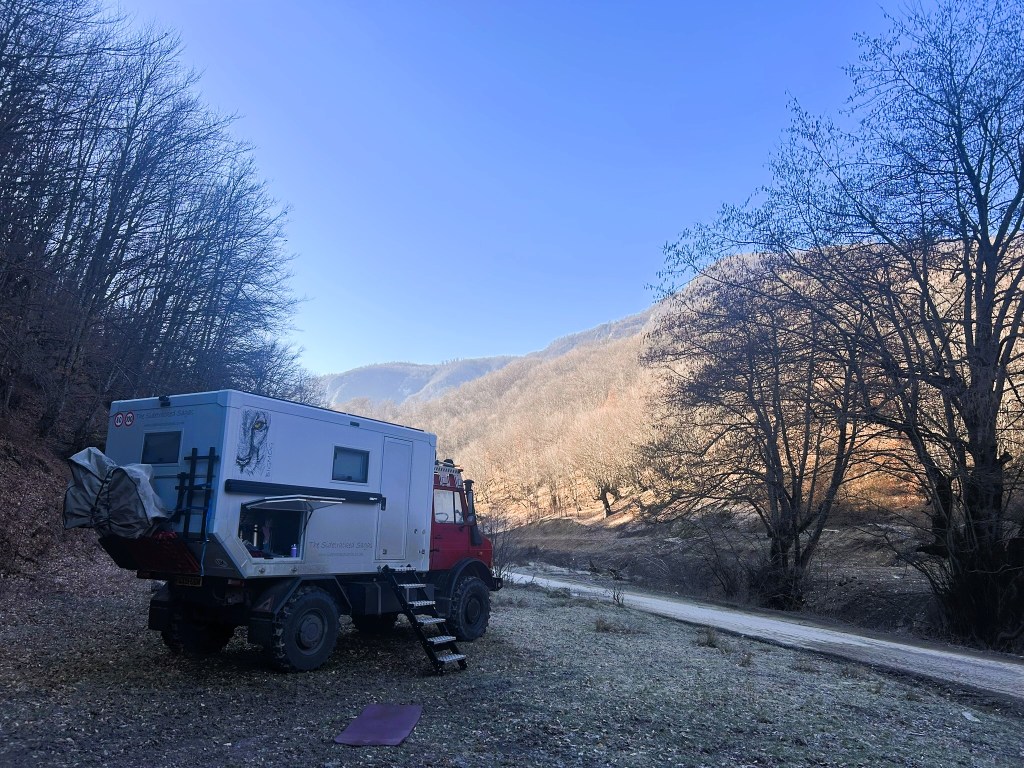 An off-road vehicle parked beside a winding dirt road, surrounded by trees and hills under a clear blue sky.