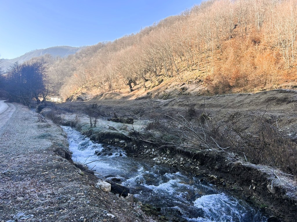 A serene river flowing through a bare landscape during early morning, with frost on the ground and trees on a slope in autumn colours.