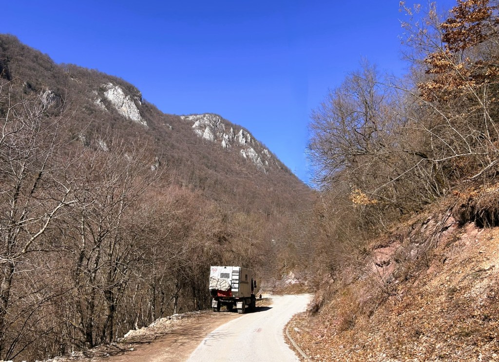 A rugged dirt road winding through barren trees and rocky mountains under a clear blue sky with a vehicle driving along the path.