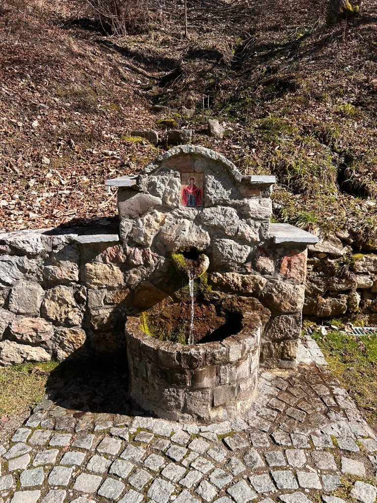 A stone water fountain with a small stone bowl, featuring a decorative arch and an icon above it, surrounded by a natural landscape and cobblestone path.