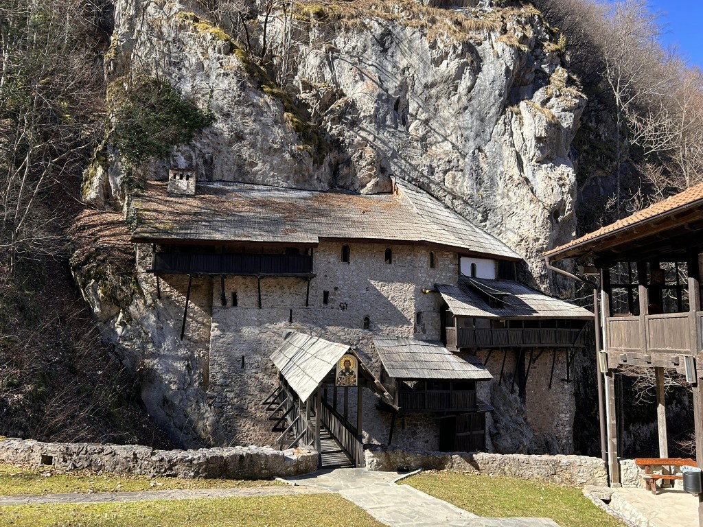 Historic building partially built into a rocky cliff, featuring a stone structure with wooden balconies and a sloping roof, surrounded by trees and natural rock formations.