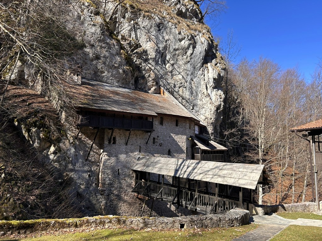 A historic building nestled against a steep rocky cliff, featuring a wooden balcony and a sloping roof, surrounded by bare trees and blue sky.