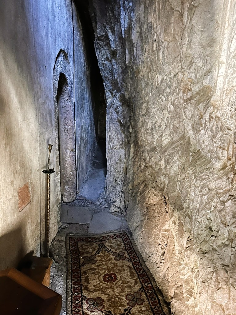 A narrow corridor with rough stone walls, featuring a patterned rug on the floor and a candlestick holder on the left side.