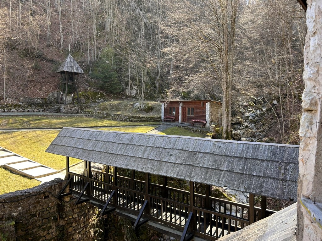 A wooden bridge with a sloped roof spans over a rocky area, leading to a small wooden building in a natural landscape with trees in the background.