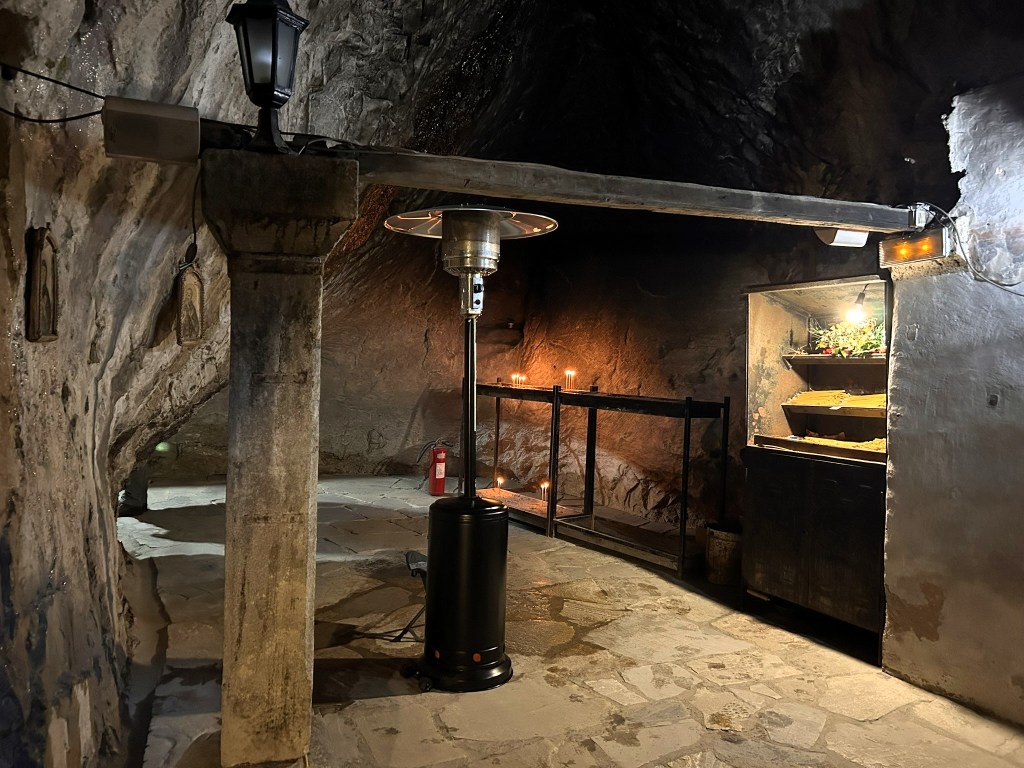 Interior of a dimly lit cave with stone walls, featuring a patio heater, shelving units, and candles, creating a unique atmospheric space.