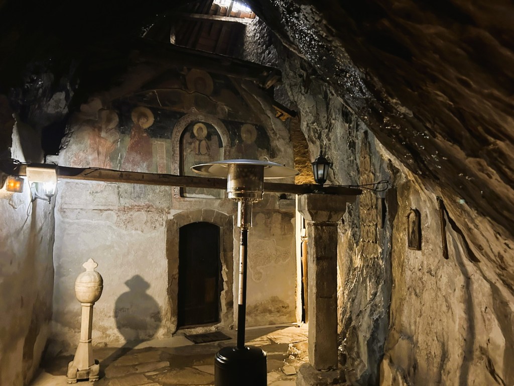 Interior of a dimly lit ancient chapel with weathered stone walls and decorative frescoes on the wall. A stone pillar stands near a doorway, while a heater and a lantern provide some light.