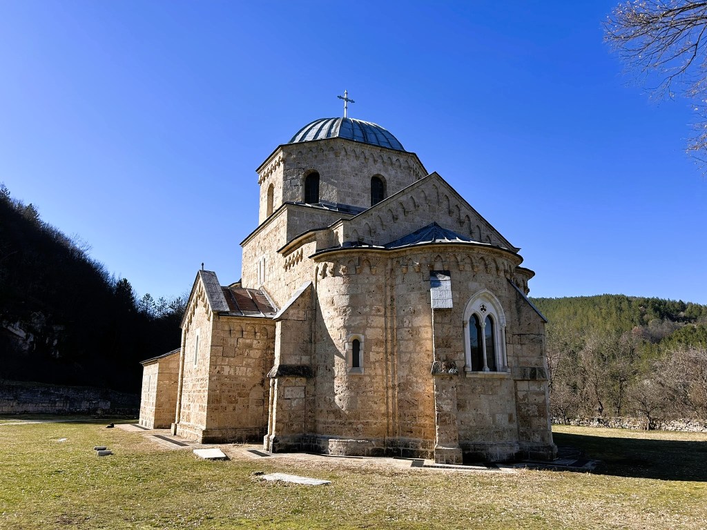 A stone church with a domed roof, set against a clear blue sky and surrounded by greenery, showcasing a blend of architectural styles.