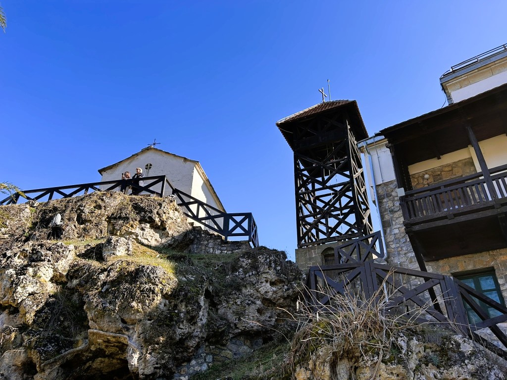 View of a rocky outcrop with a wooden railing, featuring a small church and a tall tower against a clear blue sky.