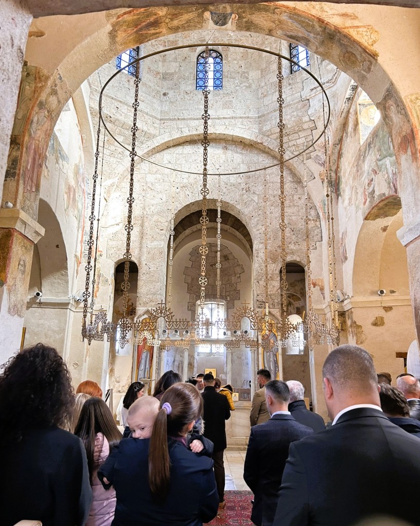 Interior of a historic church with a congregation gathered for a ceremony, featuring ornate chandeliers and religious decor.
