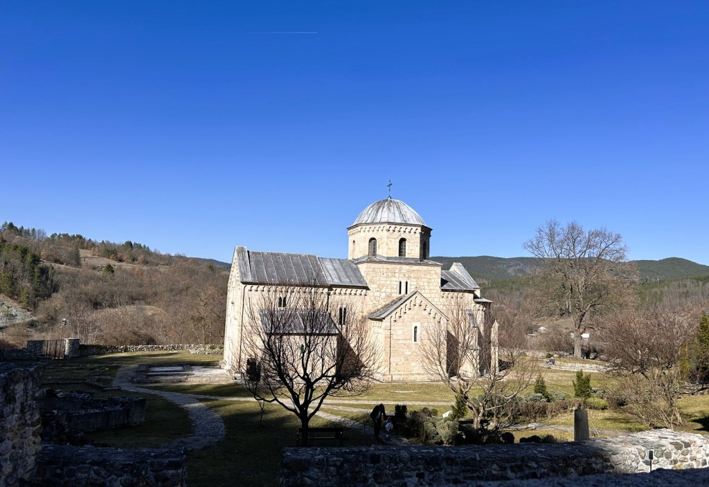 Historic stone church with a domed roof, surrounded by trees and hills under a clear blue sky.