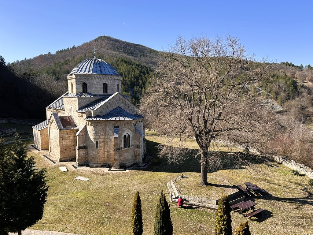A stone church with a domed roof surrounded by mountains and a tree in a grassy area, with benches and a person seated nearby.