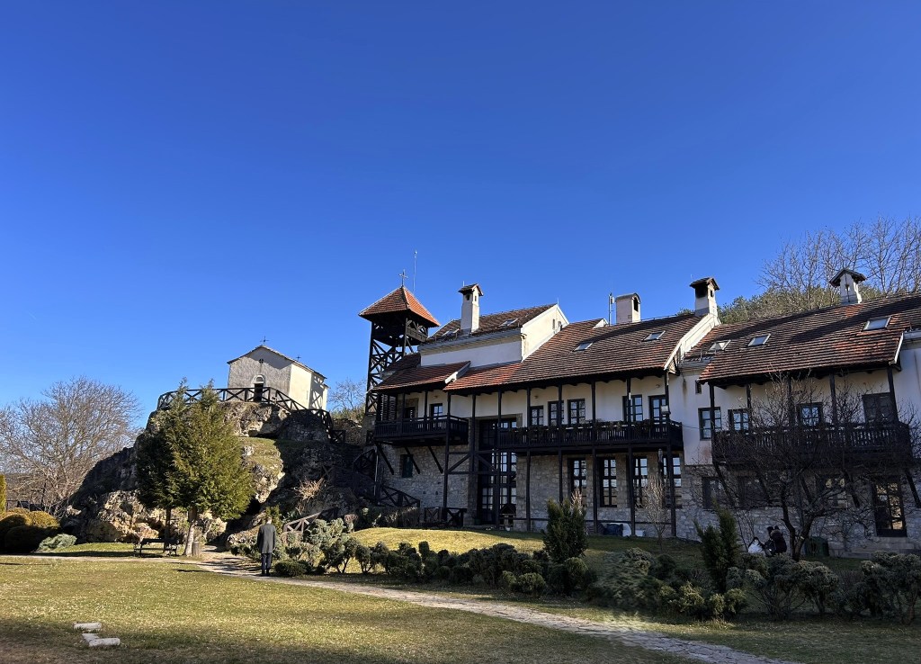 A historic building with a stone base and wooden balconies set against a clear blue sky, surrounded by landscaped gardens and trees.