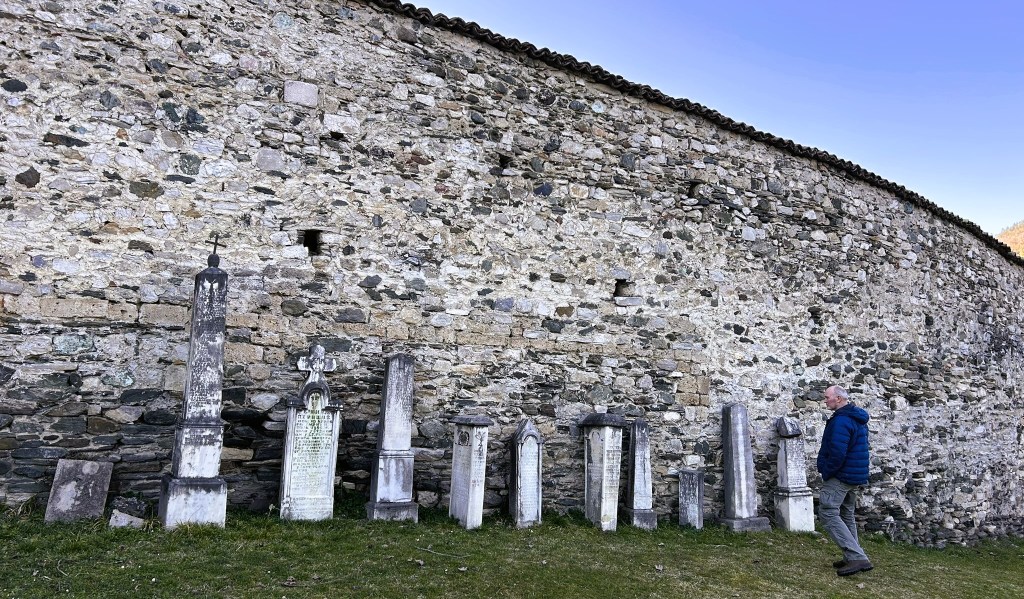 A person stands beside a row of gravestones against a stone wall, with a clear sky in the background.