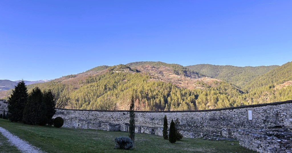 A scenic view of a mountainous landscape with lush greenery and a clear blue sky, featuring a stone wall in the foreground.