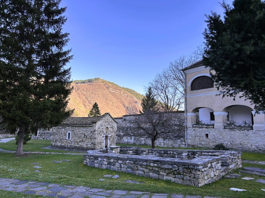 A peaceful landscape featuring a stone structure surrounded by grass, trees, and mountains in the background under a clear blue sky.