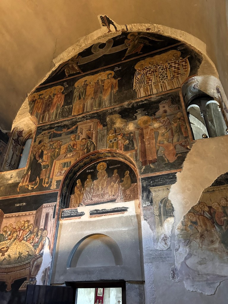 A detailed view of ancient frescoes on a church wall, featuring religious figures and scenes. The artwork includes numerous saints and biblical imagery, with a sense of historical significance.