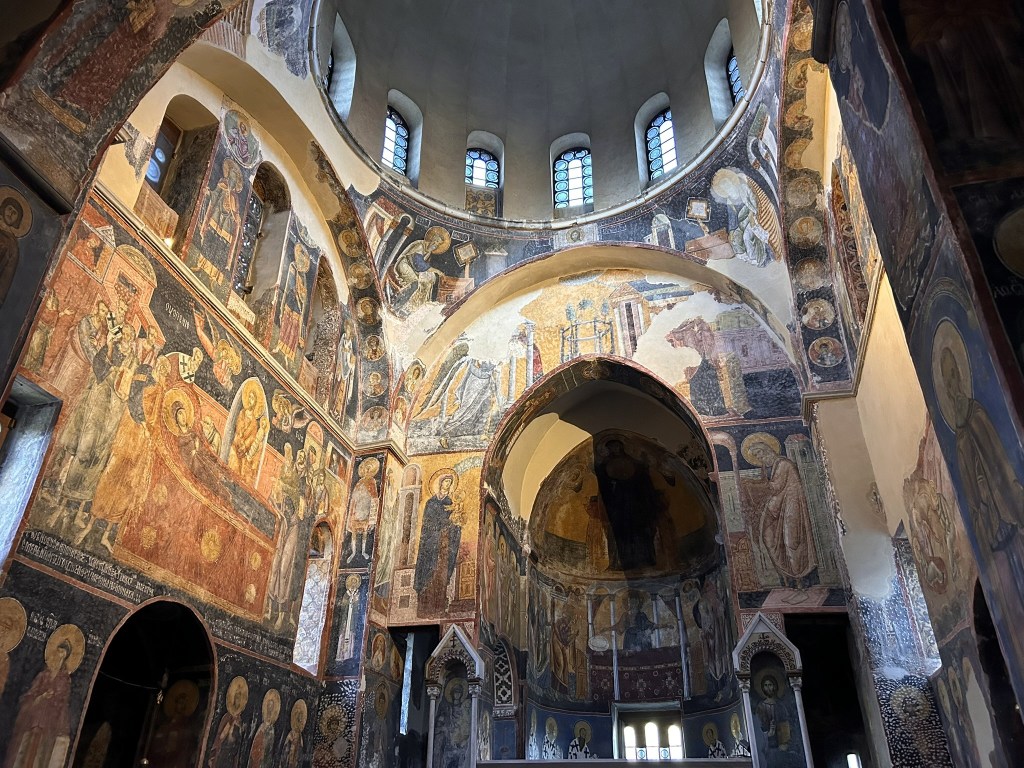 Interior of an ancient church with ornate frescoes depicting religious scenes on the walls and a high arching ceiling.